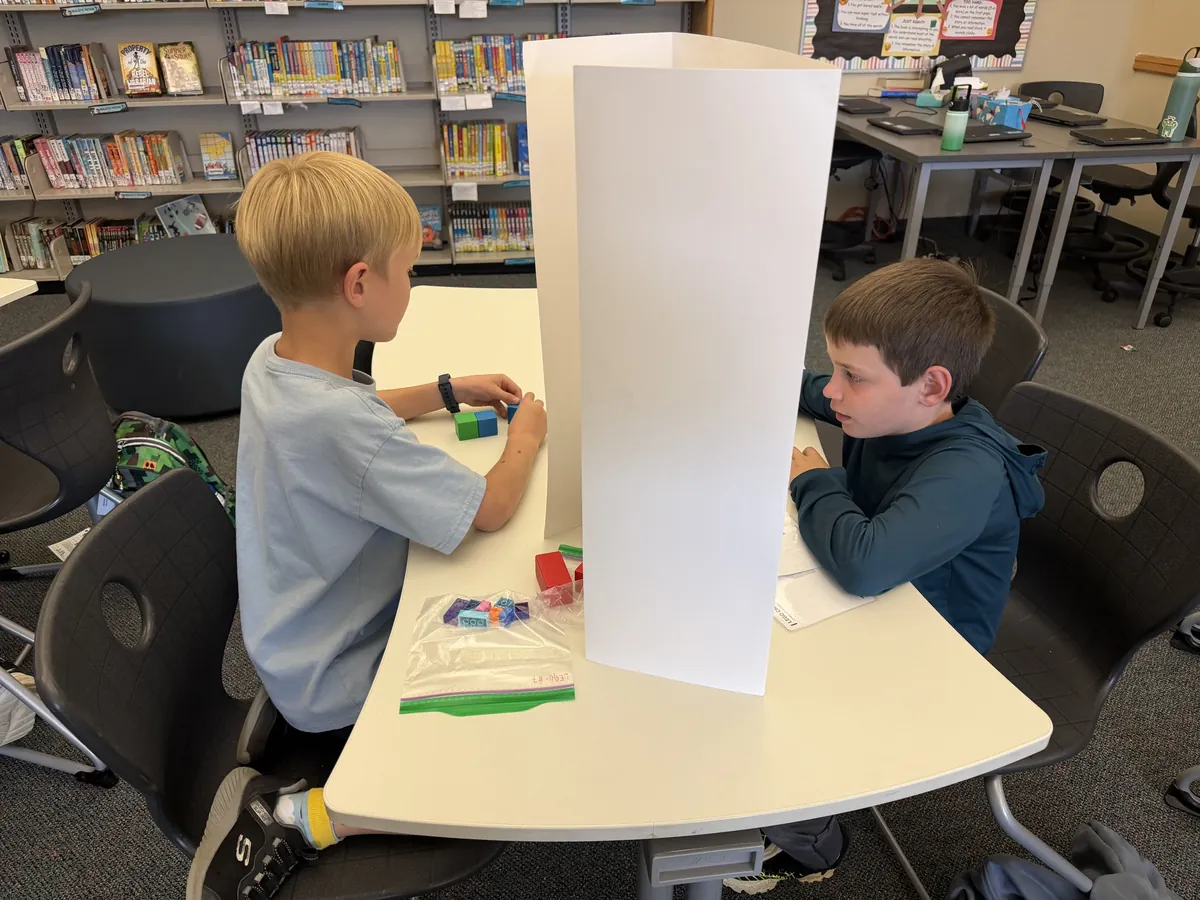 Two students at a table separated by a white wall, one building with blocks while the other gives instructions